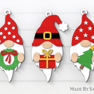 May include: Three wooden Christmas gnome ornaments with white beards and red hats. The gnome on the left has a red and white polka dot hat and is holding a green wreath. The gnome in the middle has a red hat with a black belt and is holding a red gift box with a yellow bow. The gnome on the right has a red hat with white stars and is holding a green Christmas tree.