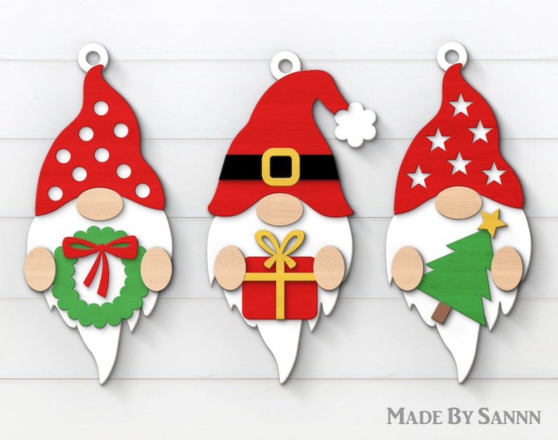 May include: Three wooden Christmas gnome ornaments with white beards and red hats. The gnome on the left has a red and white polka dot hat and is holding a green wreath. The gnome in the middle has a red hat with a black belt and is holding a red gift box with a yellow bow. The gnome on the right has a red hat with white stars and is holding a green Christmas tree.