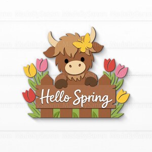 Peut inclure: D&eacute;coration printani&egrave;re en bois avec une vache des Highlands de dessin anim&eacute;, un papillon jaune, et une cl&ocirc;ture marron avec l'inscription "Hello Spring" en blanc, entour&eacute;e de tulipes color&eacute;es.