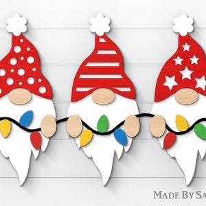 Peut inclure: Décorations de Noël en bois représentant des gnomes avec des chapeaux rouges à pois blancs, rayures et étoiles. Chaque gnome a un nez en bois, une barbe blanche et tient une guirlande lumineuse colorée. Le texte "MADE BY SANNN" est visible.