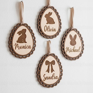 May include: Four wooden Easter egg decorations with scalloped edges and brown ribbon hangers. Each egg features a different design: a bunny, a bow, and two different bunny silhouettes. Names are engraved: Monica, Olivia, Michael, and Sandra.