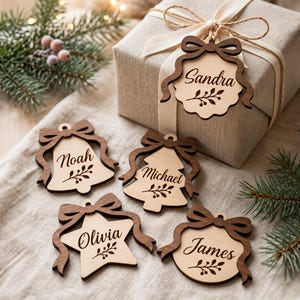 Op de afbeelding: Vijf houten cadeaulabels met donkerbruine randen en lichte houten centra. Elk label heeft een strik, een takje-illustratie en een in script gegraveerde naam: Sandra, Noah, Michael, Olivia en James. Eén label zit aan een doos.