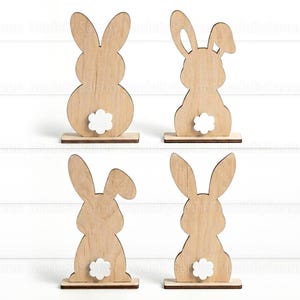 May include: Four wooden bunny ornaments with a light brown finish and white flower-shaped tails. Each bunny is mounted on a small wooden base. The Easter decorations are arranged on a white surface.