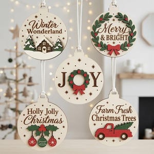 Op de afbeelding: Vijf ronde houten ornamenten met witte oppervlakken en ophangkoorden. De ontwerpen bevatten een huis met bomen en de tekst Winter Wonderland, een krans met Merry & Bright, een krans met het woord JOY, hulst met Holly Jolly Christmas en een rode vrachtwagen met Farm Fresh Christmas Trees.