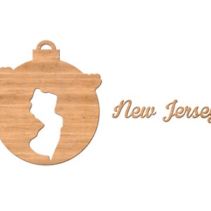 Pode incluir: Enfeite de Natal de madeira em forma de c&iacute;rculo com o contorno do estado de Nova Jersey recortado. O enfeite &eacute; marrom e tem uma textura de gr&atilde;o de madeira. O texto "New Jersey" est&aacute; escrito em it&aacute;lico abaixo do enfeite.