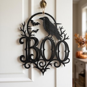 Puede incluir: Un adorno de pared de madera negra cortado con l&aacute;ser que presenta la palabra BOO en una fuente decorativa. El dise&ntilde;o incluye un cuervo posado sobre las letras, dos peque&ntilde;os murci&eacute;lagos volando y un marco circular con ramas de &aacute;rbol.