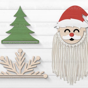 Può includere: Un set di tre decorazioni natalizie in legno: un albero di Natale verde, un fiocco di neve bianco e una testa di Babbo Natale con una barba di lana bianca.