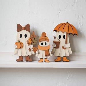 Puede incluir: Tres figuras de fantasmas de madera plana en fila. El fantasma de la izquierda lleva un lazo marr&oacute;n, sostiene una calabaza y hojas de oto&ntilde;o, y usa botas marrones. El fantasma central lleva un gorro y bufanda. El de la derecha sostiene un caf&eacute;.