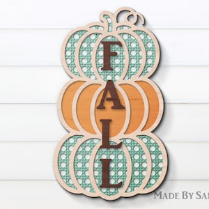 Puede incluir: Un recorte de madera de tres calabazas apiladas con la palabra "FALL" en letras marrones. Las calabazas están decoradas con un patrón de cuadros verdes y blancos.