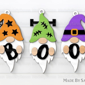 May include: Three wooden Halloween gnomes with orange, green, and purple hats. The gnomes spell out "BOO" with black letters. The gnome with the orange hat has a spider web and stars on his hat. The gnome with the green hat has stitches on his hat. The gnome with the purple hat has a spider on his hat.