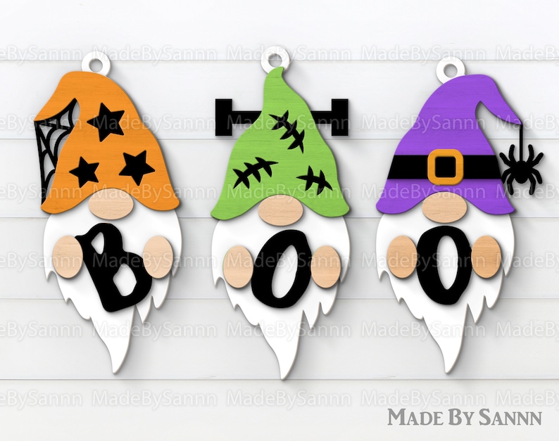 May include: Three wooden Halloween gnomes with orange, green, and purple hats. The gnomes spell out "BOO" with black letters. The gnome with the orange hat has a spider web and stars on his hat. The gnome with the green hat has stitches on his hat. The gnome with the purple hat has a spider on his hat.