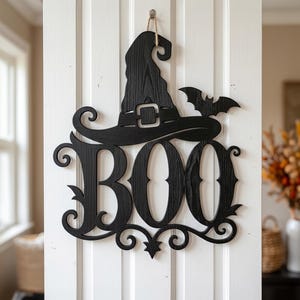 Puede incluir: Un adorno de pared de madera negra con la palabra BOO en una fuente estilizada, rematado con un sombrero de bruja y la silueta de un murci&eacute;lago. La pieza muestra una veta de madera visible y cuelga de un gancho mediante un cordel fino.