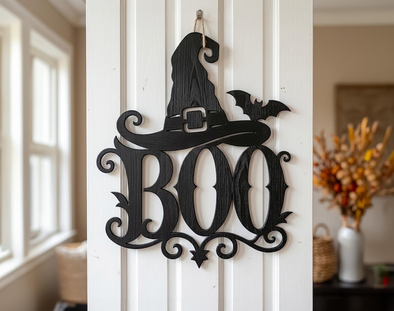 Puede incluir: Un adorno de pared de madera negra con la palabra BOO en una fuente estilizada, rematado con un sombrero de bruja y la silueta de un murci&eacute;lago. La pieza muestra una veta de madera visible y cuelga de un gancho mediante un cordel fino.