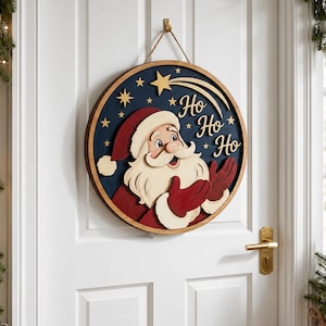 May include: A circular wooden wall sign featuring a layered design of Santa Claus in a red suit and hat against a dark blue background. The sign includes gold-colored stars, a shooting star, and the text "Ho Ho Ho" in a script font, suspended by a rope.