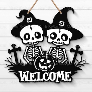 Op de afbeelding: Zwarte en witte Halloween-deurhanger met twee skeletfiguren die toverhoeden dragen en een jack-o-lantern vasthouden. Het bordje staat "WELCOME".