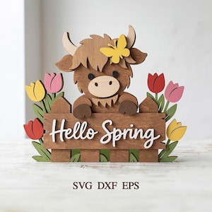 Puede incluir: Decoración de primavera de madera con una vaca de las Tierras Altas asomándose por encima de una valla con las palabras "Hello Spring". La vaca tiene pelaje marrón, una mariposa amarilla y está rodeada de tulipanes coloridos. Un encantador acento estacional.