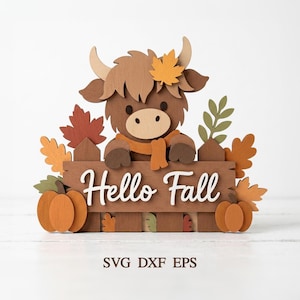 May include: A layered wooden cutout decoration featuring a brown highland cow with a yellow leaf on its head, standing behind a brown fence with the white text Hello Fall. The scene includes orange pumpkins and various autumn leaves in shades of red, orange, and green.