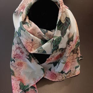 May include: A lightweight scarf featuring a floral pattern with large, soft pink and peach-colored dahlia blossoms interspersed with deep green leaves against a pale, off-white background. The fabric is draped to show the intricate botanical print.