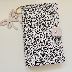 May include: A closed notebook with a leopard print cover in black and white, featuring a pink glittery clasp and a matching bow-shaped keyring. The notebook is closed and has a gold-coloured ring.