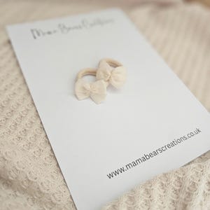 May include: Two beige baby hair ties with bow accents rest on a white card. The card has the text "Mama Bears Creations" and the website address "www.mamabearscreations.co.uk". The background is a soft, textured fabric.