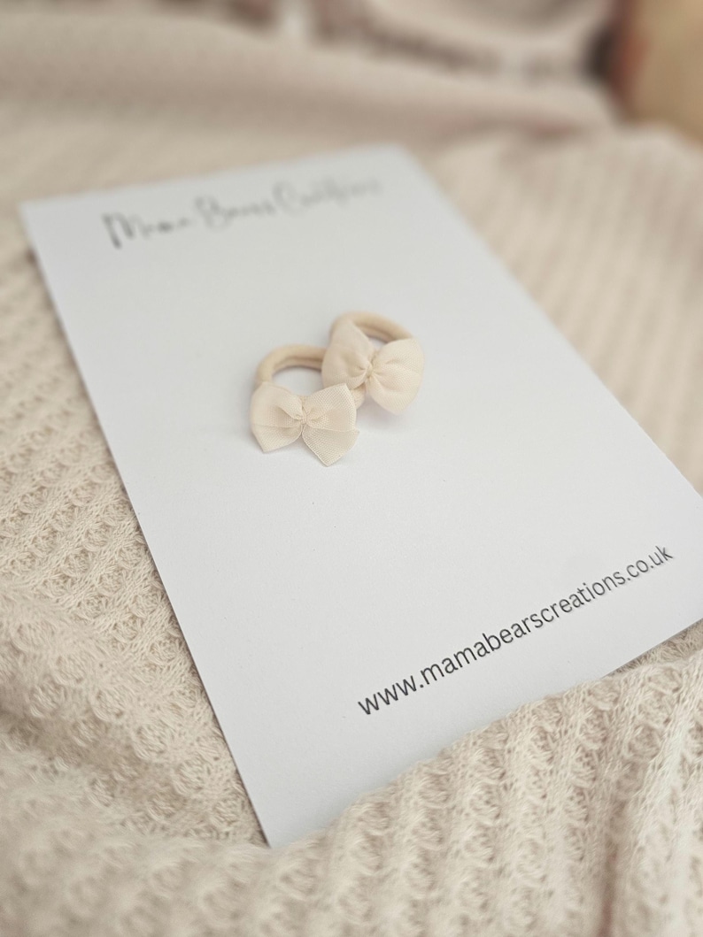 May include: Two beige baby hair ties with bow accents rest on a white card. The card has the text "Mama Bears Creations" and the website address "www.mamabearscreations.co.uk". The background is a soft, textured fabric.