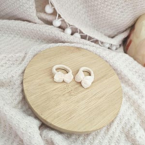 May include: Two small, light pink bow hair ties rest on a round, light brown wooden surface. The background features a soft, white, textured blanket. The hair accessories are delicate and feminine, perfect for adding a touch of charm to any hairstyle.