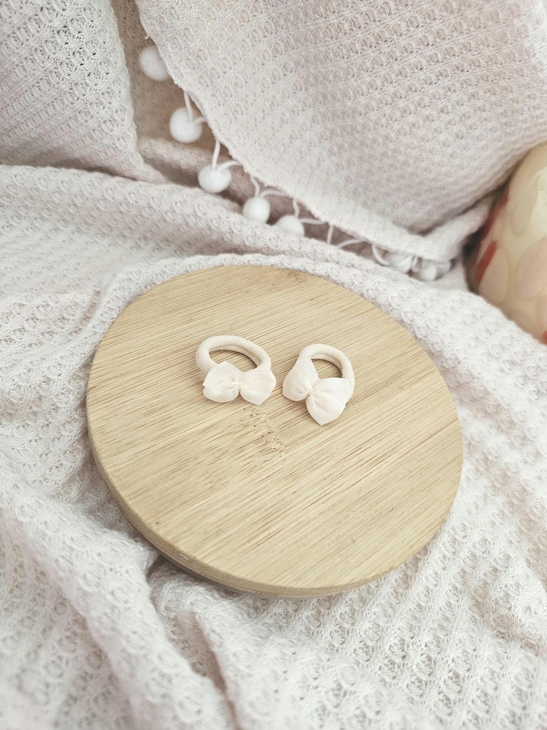 May include: Two small, light pink bow hair ties rest on a round, light brown wooden surface. The background features a soft, white, textured blanket. The hair accessories are delicate and feminine, perfect for adding a touch of charm to any hairstyle.