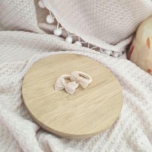 May include: Two light pink bow hair ties rest on a round wooden surface. The bows are small and delicate, with a soft, matte finish. The wooden surface is light-colored and has a natural grain. The background features a textured white blanket.