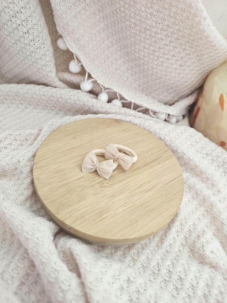 May include: Two light pink bow hair ties rest on a round wooden surface. The bows are small and delicate, with a soft, matte finish. The wooden surface is light-colored and has a natural grain. The background features a textured white blanket.