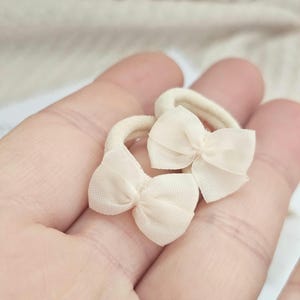 May include: Two small, cream-colored hair ties with delicate bow accents. The bows are made of a sheer fabric, adding a touch of elegance. The hair ties are held in a hand, suggesting their size and use. Perfect for adding a subtle, feminine touch to any hairstyle.