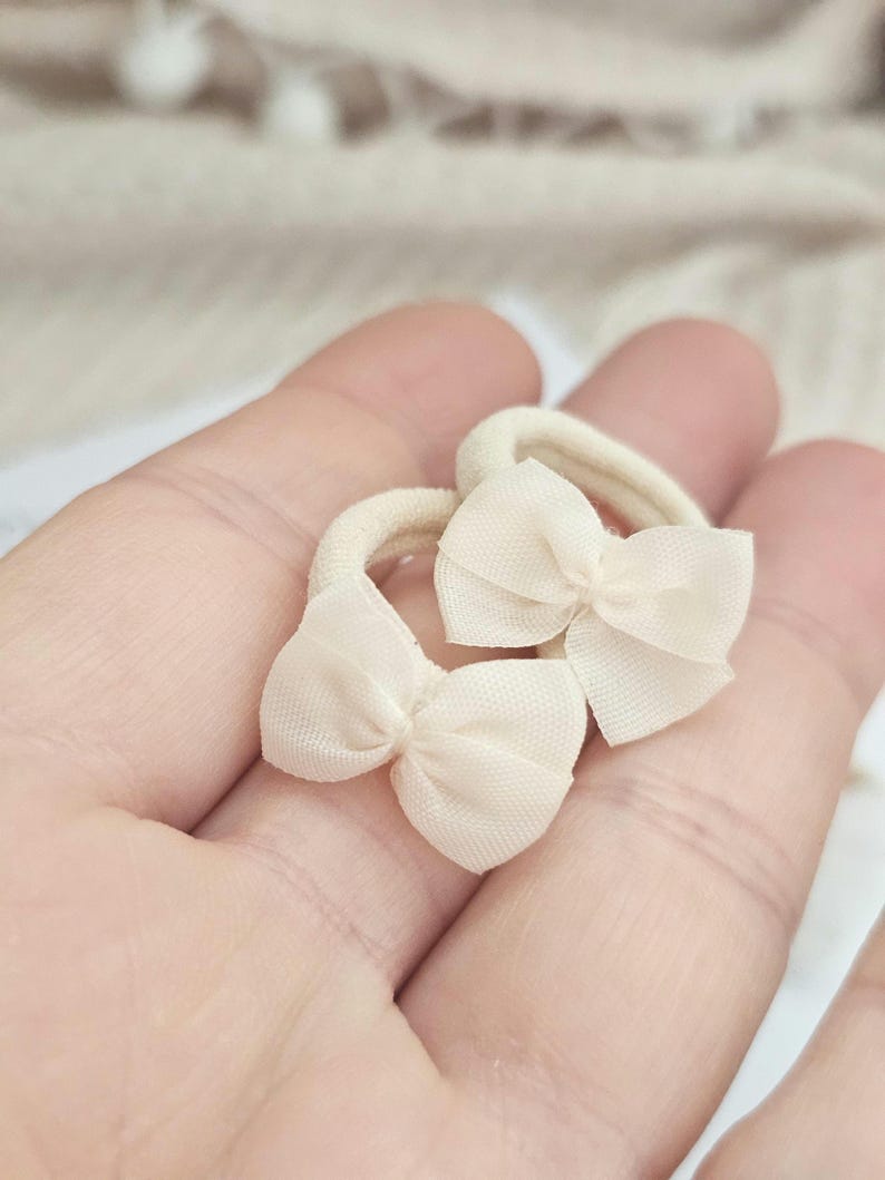 May include: Two small, cream-colored hair ties with delicate bow accents. The bows are made of a sheer fabric, adding a touch of elegance. The hair ties are held in a hand, suggesting their size and use. Perfect for adding a subtle, feminine touch to any hairstyle.