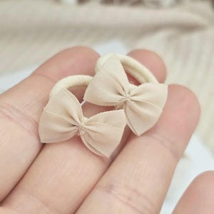 May include: Two beige hair ties with small bow accents. The bows are made of a sheer fabric, and the elastic bands are a matching beige color. The hair accessories are shown against a soft, neutral background.