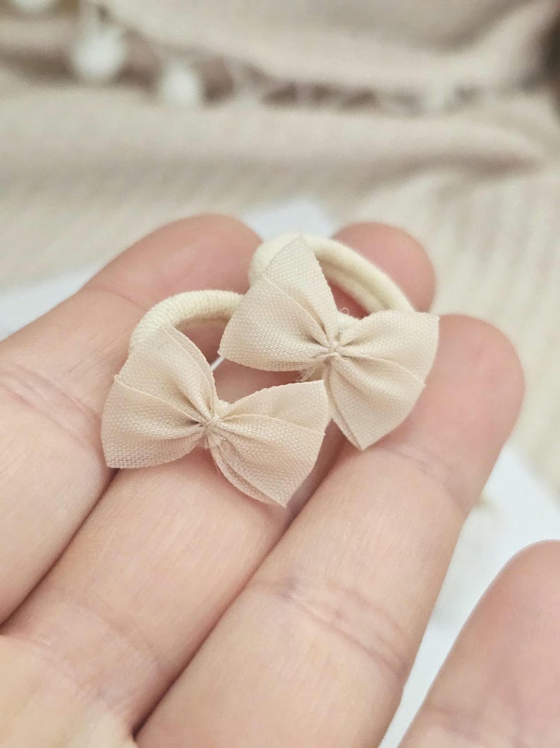 May include: Two beige hair ties with small bow accents. The bows are made of a sheer fabric, and the elastic bands are a matching beige color. The hair accessories are shown against a soft, neutral background.