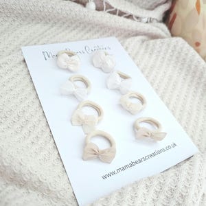 May include: A collection of eight hair ties with small bow accents in shades of white and beige. The hair ties are arranged on a white card with the text "Mama Bears Creations" and the website address www.mamabearscreations.co.uk.