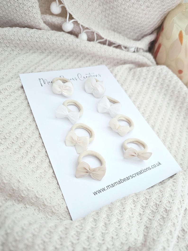 May include: A collection of eight hair ties with small bow accents in shades of white and beige. The hair ties are arranged on a white card with the text "Mama Bears Creations" and the website address www.mamabearscreations.co.uk.