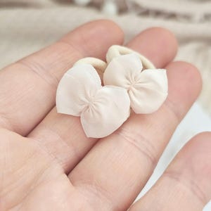 May include: Two small, light beige flower-shaped hair accessories. Each accessory features a delicate, petal-like design made of a sheer fabric, attached to a beige elastic band. The accessories are designed for hair styling.