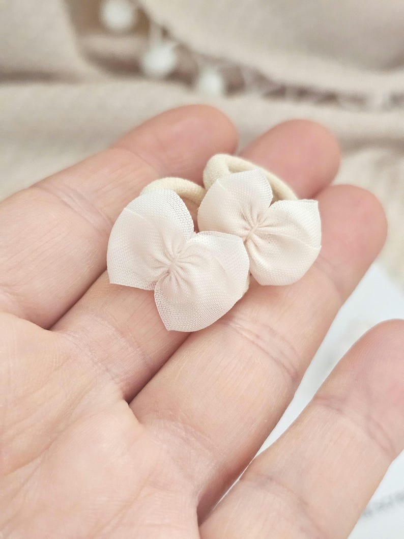 May include: Two small, light beige flower-shaped hair accessories. Each accessory features a delicate, petal-like design made of a sheer fabric, attached to a beige elastic band. The accessories are designed for hair styling.