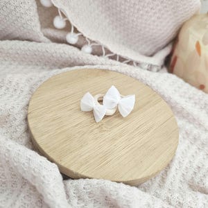 May include: Two small white bow hair clips rest on a light wooden round surface. The clips are made of fabric and are tied in a bow shape. The wooden surface sits on a soft, textured, off-white blanket.