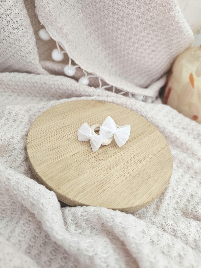 May include: Two small white bow hair clips rest on a light wooden round surface. The clips are made of fabric and are tied in a bow shape. The wooden surface sits on a soft, textured, off-white blanket.