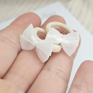 May include: Two small, cream-colored hair ties with white bow accents. The bows are made of a sheer fabric, and the hair ties are held in a person's hand. These accessories are suitable for babies, toddlers, or anyone who enjoys a delicate hair accessory.