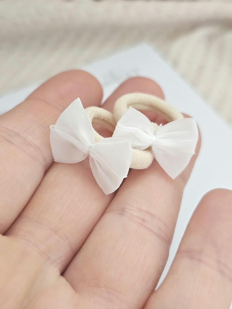 May include: Two small, cream-colored hair ties with white bow accents. The bows are made of a sheer fabric, and the hair ties are held in a person's hand. These accessories are suitable for babies, toddlers, or anyone who enjoys a delicate hair accessory.
