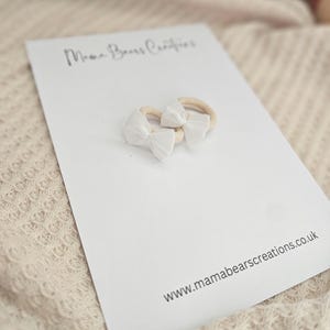 May include: Two white bow headbands on a white card with the text "Mama Bears Creations" and "www.mamabearscreations.co.uk". The headbands have a beige elastic band.