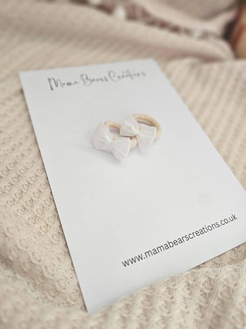 May include: Two white bow headbands on a white card with the text "Mama Bears Creations" and "www.mamabearscreations.co.uk". The headbands have a beige elastic band.