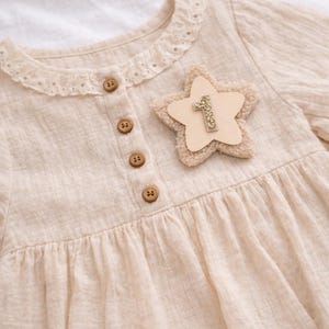 Op de afbeelding: Crèmekleurige babyjurk met een stervormige applicatie. De jurk heeft een ronde, met kant afgewerkte kraag, houten knopen en een gerimpelde rok. De sterapplicatie heeft een pluizige rand en het goudkleurige nummer "1" in het midden.