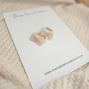 May include: Two beige bow hair ties on a white card with the text "Mama Bears Creations" and the website address www.mamabearscreations.co.uk. The hair ties are made of a soft material and are in a neutral color.