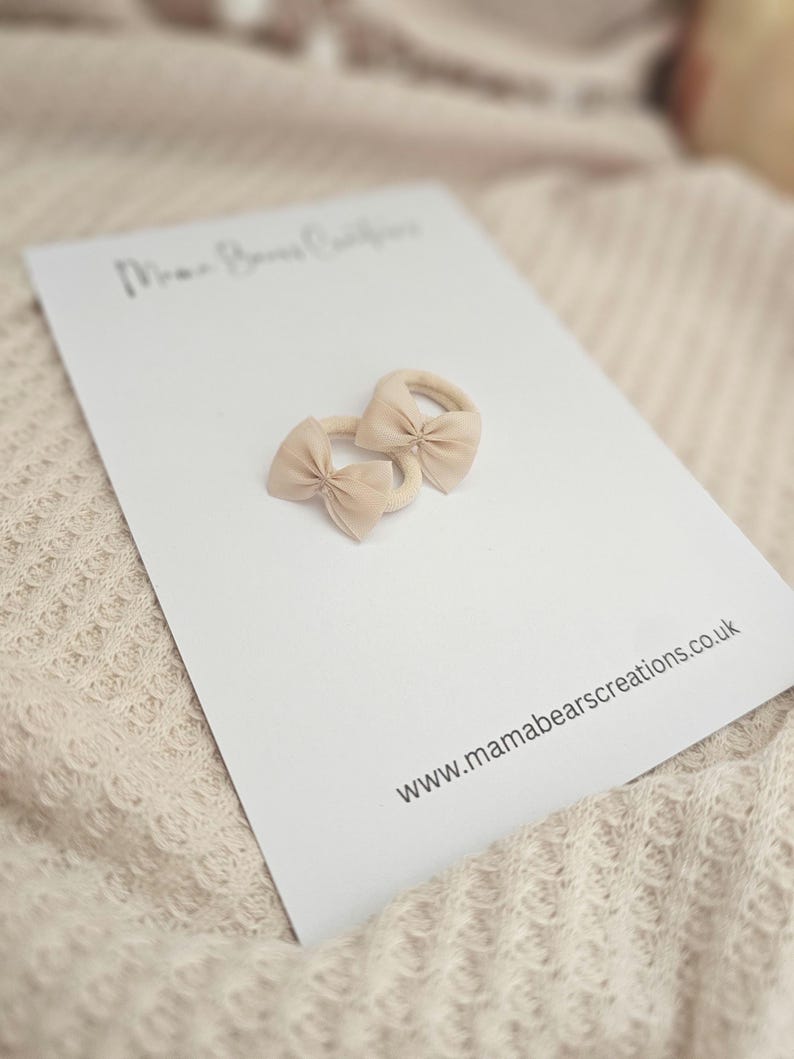 May include: Two beige bow hair ties on a white card with the text "Mama Bears Creations" and the website address www.mamabearscreations.co.uk. The hair ties are made of a soft material and are in a neutral color.