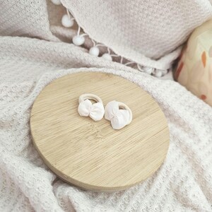 May include: Two white hair ties with small pink bows rest on a round wooden surface. The ties are set against a backdrop of a textured, off-white blanket and a decorative ceramic item.