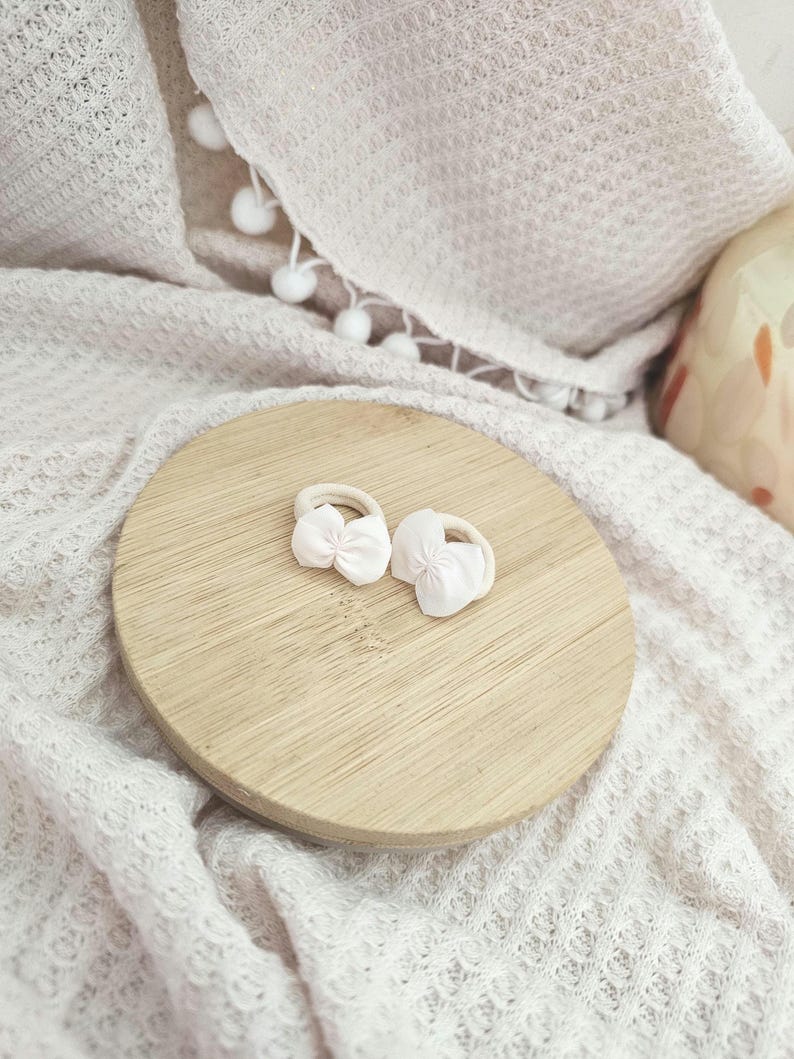 May include: Two white hair ties with small pink bows rest on a round wooden surface. The ties are set against a backdrop of a textured, off-white blanket and a decorative ceramic item.