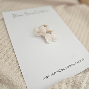 May include: Two small, light pink bow hair ties on a white card. The card has the text "Mama Bears Creations" and the website address www.mamabearscreations.co.uk. The background is a soft, neutral color.