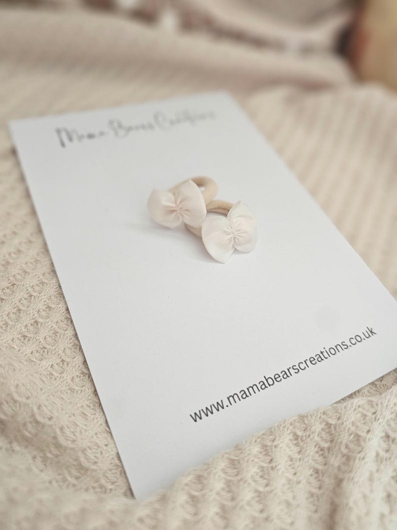 May include: Two small, light pink bow hair ties on a white card. The card has the text "Mama Bears Creations" and the website address www.mamabearscreations.co.uk. The background is a soft, neutral color.